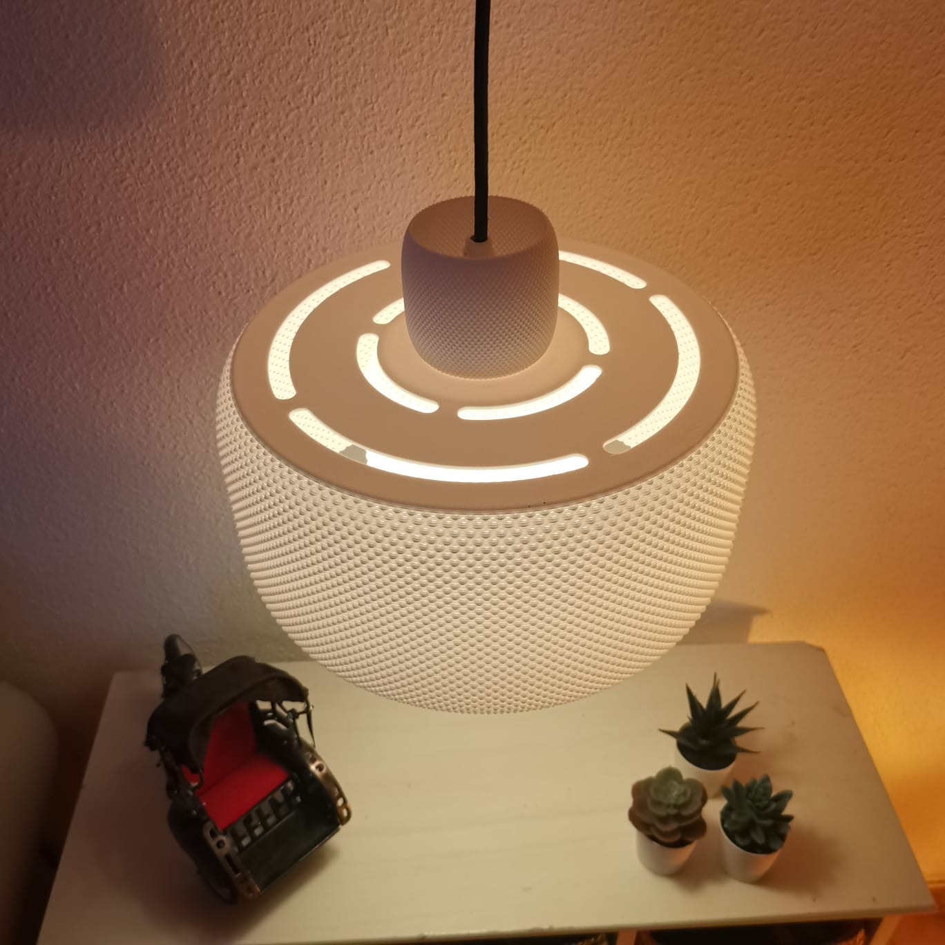 Modern pendant light with a textured design on a white surface, accompanied by small potted plants.