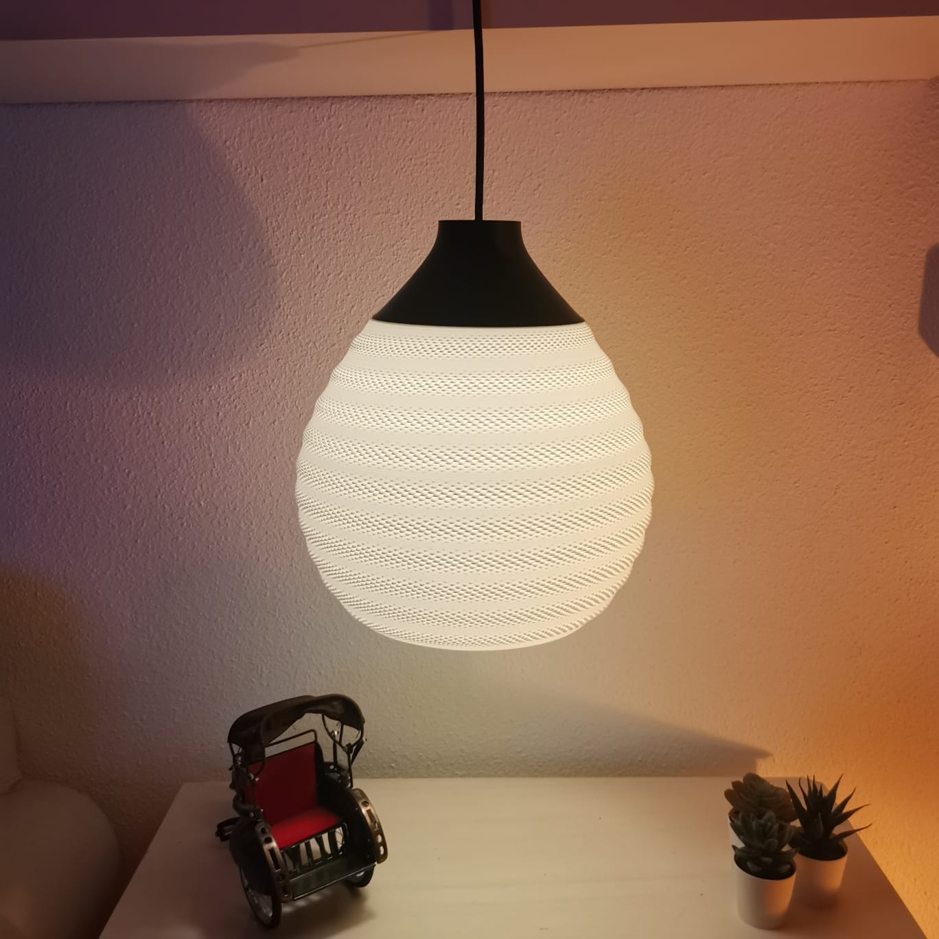 White textured pendant light on a white surface with small plants and a toy car.