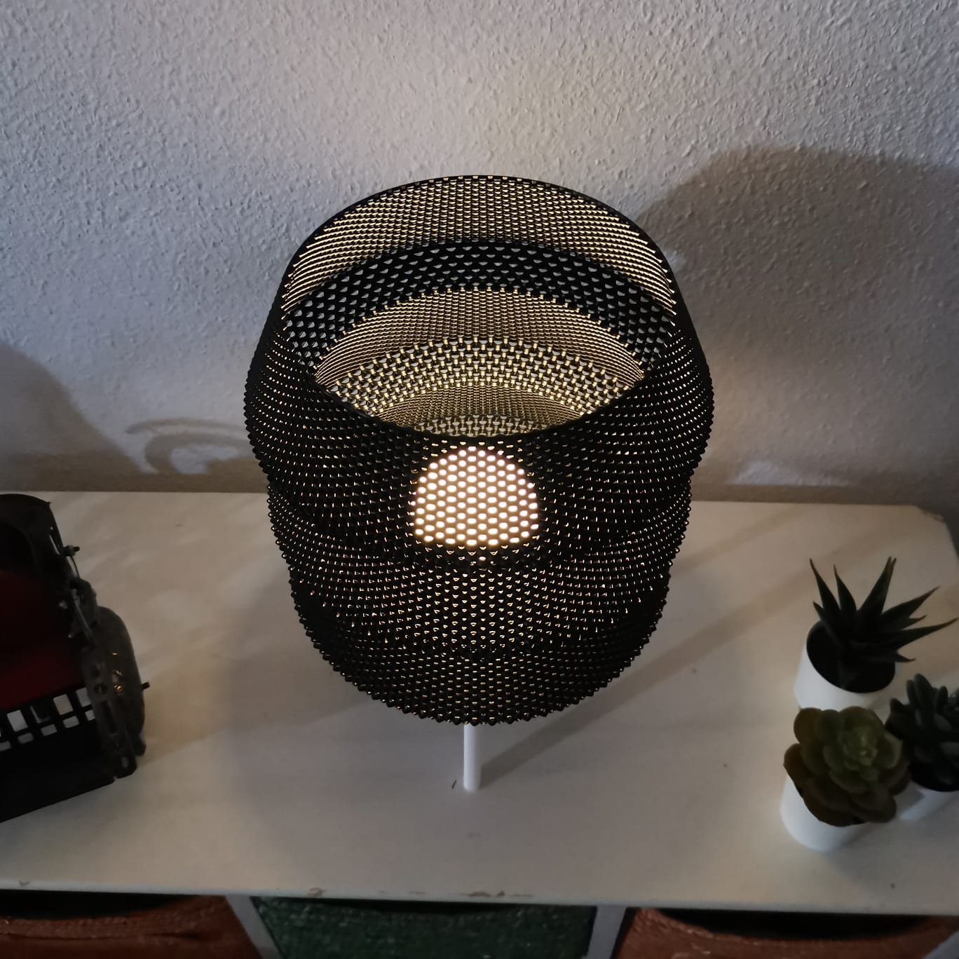 Black mesh table lamp on a white surface with plants in the background