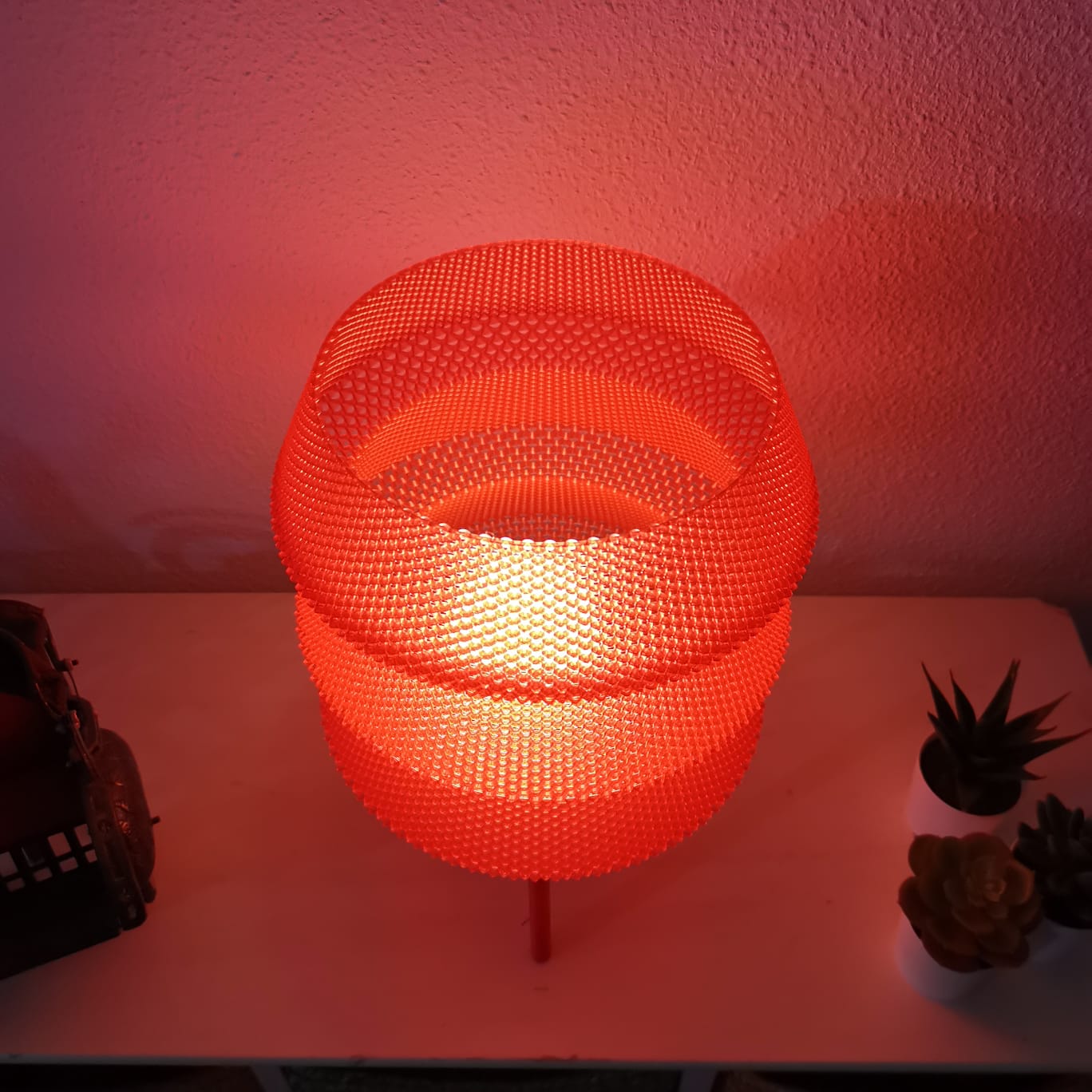 Round woven lamp with a warm glow on a surface against a pinkish wall.