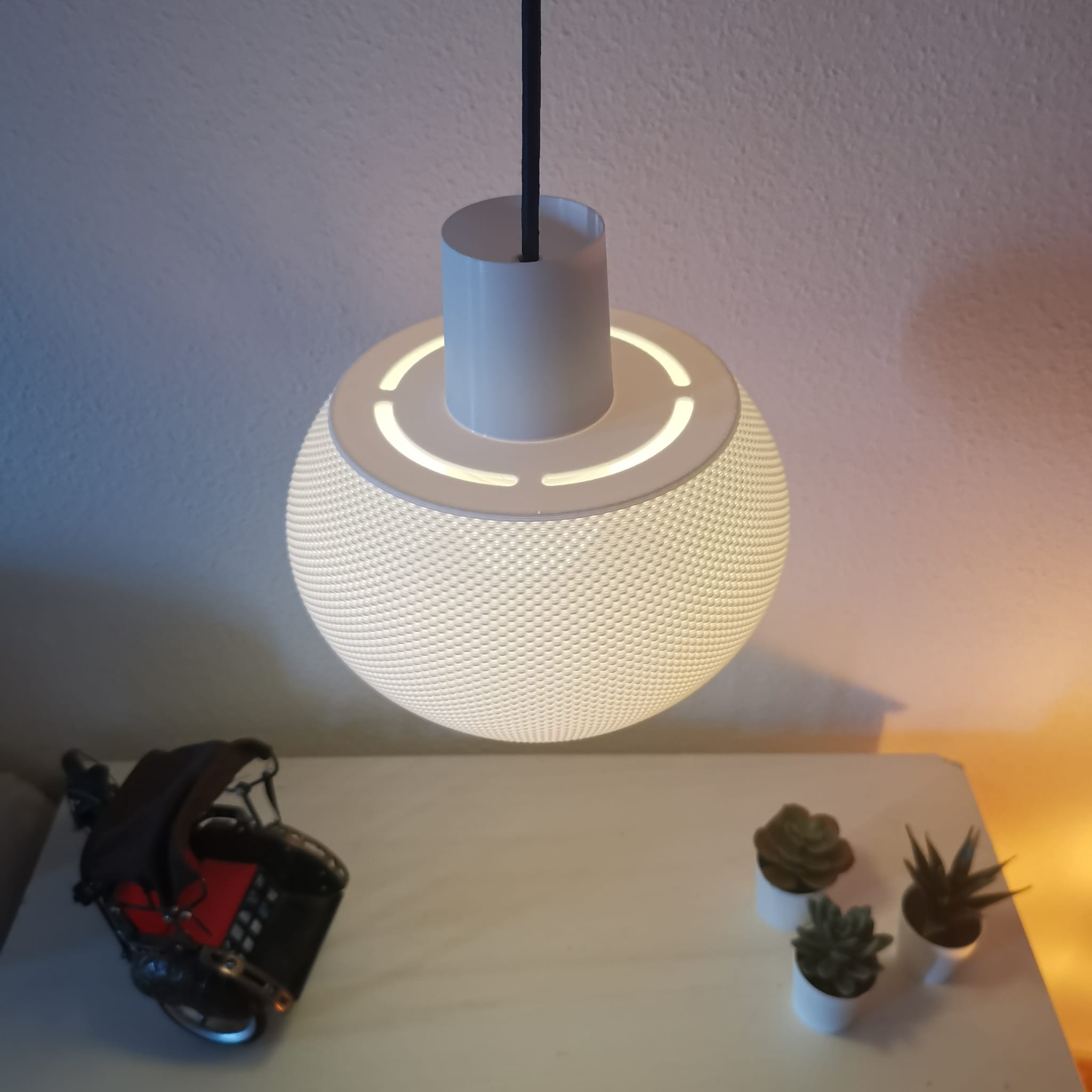 Modern pendant light with a textured design on a white surface next to small potted plants.