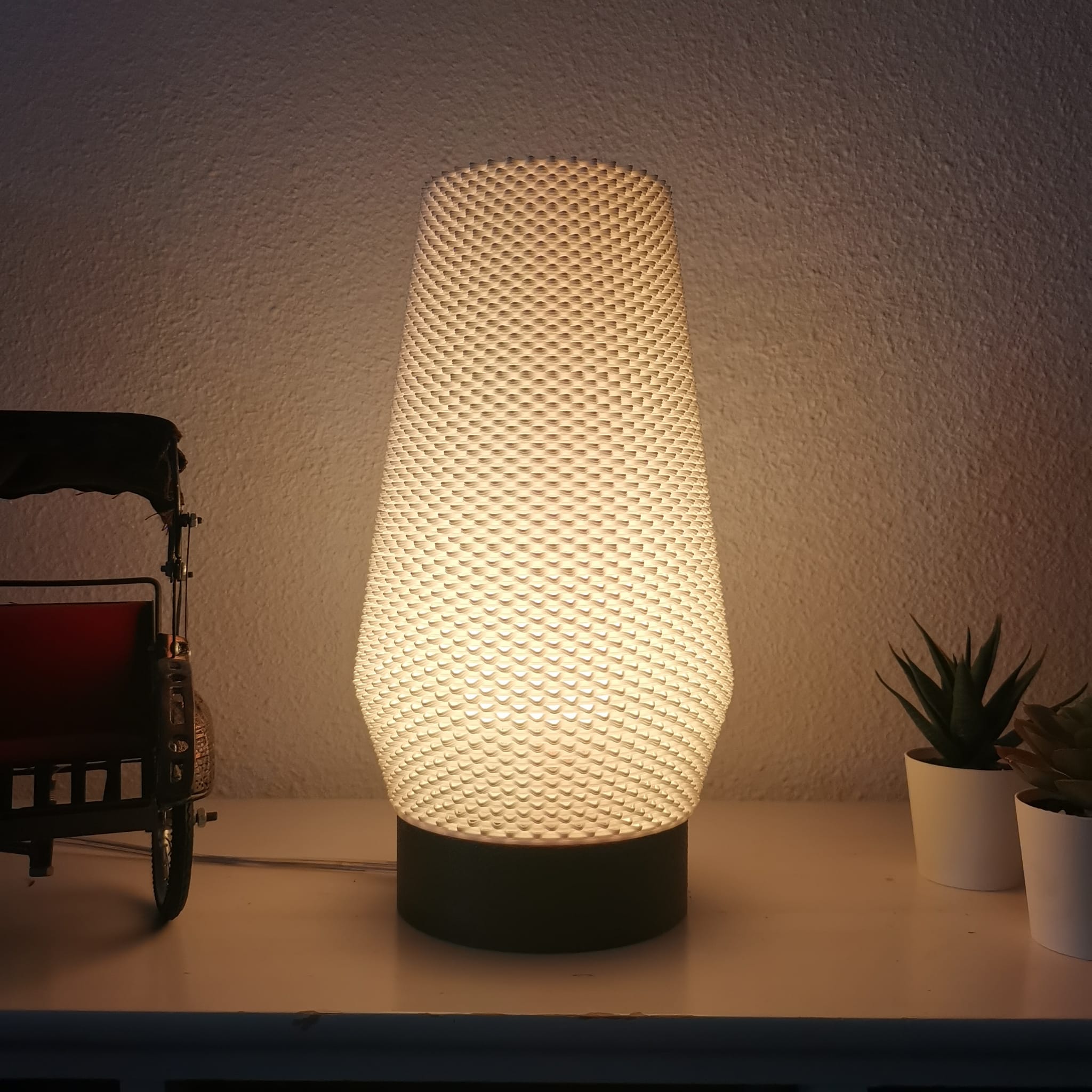 Textured table lamp on a surface with a warm glow, next to a small decorative cart and plants.