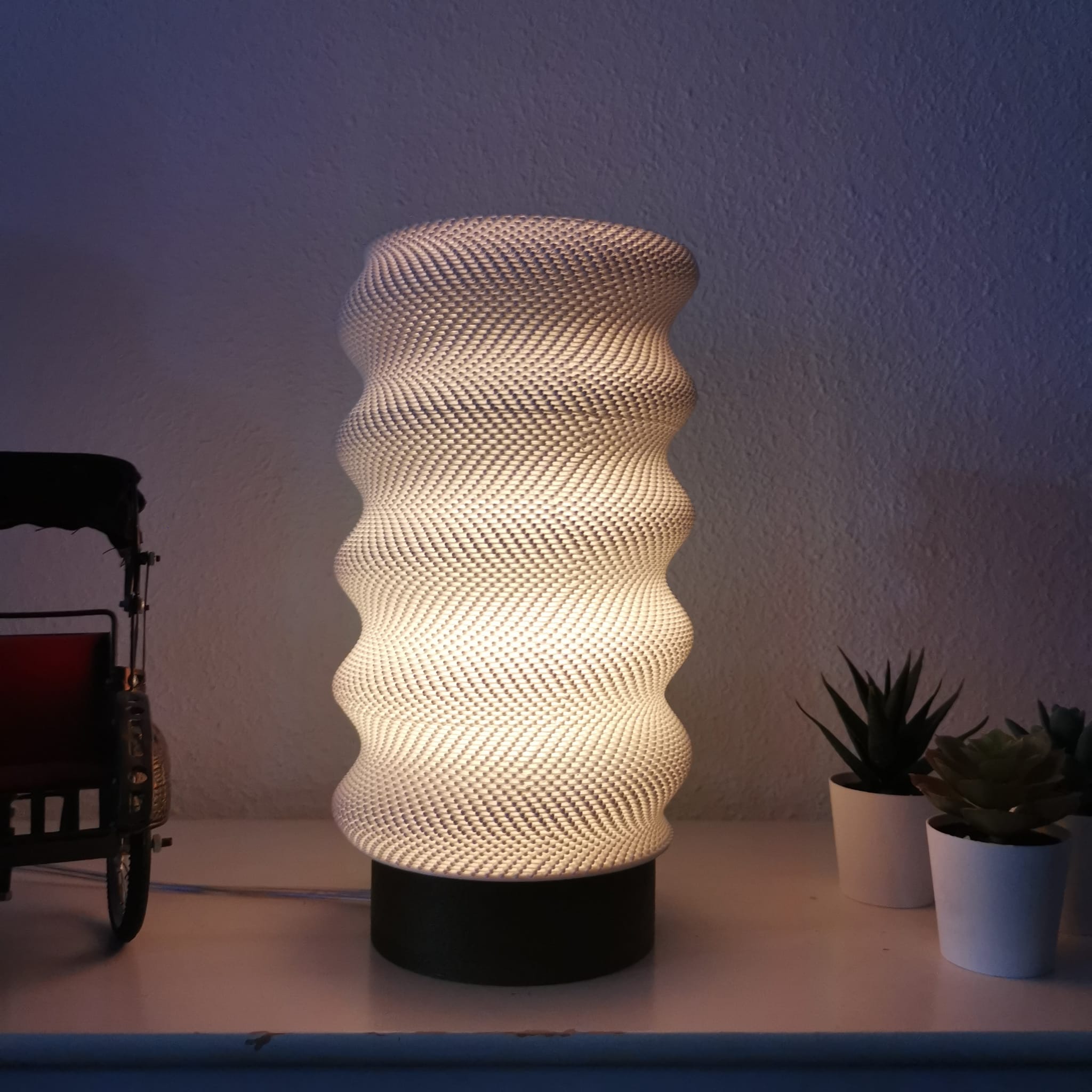 Decorative lamp with a textured design on a table with plants and a cart in the background.
