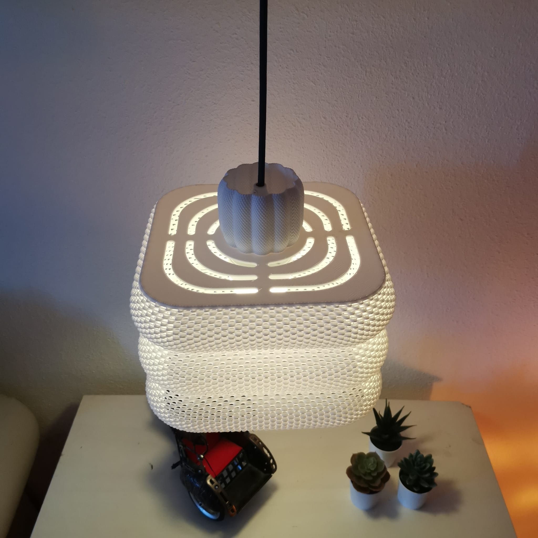 Square lamp with a textured design on a table with small plants.
