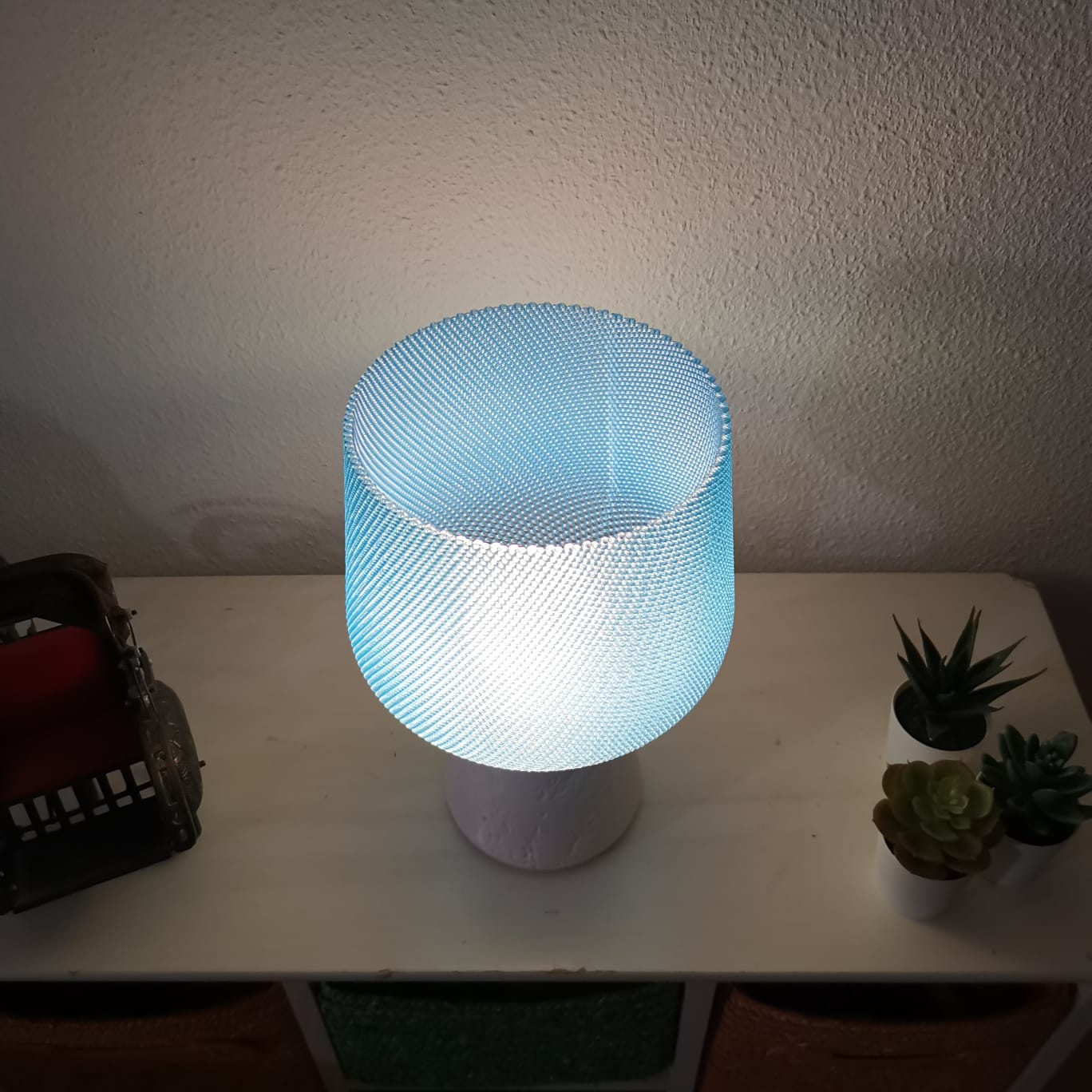 Table lamp with a textured blue shade on a white surface, with plants in the background.