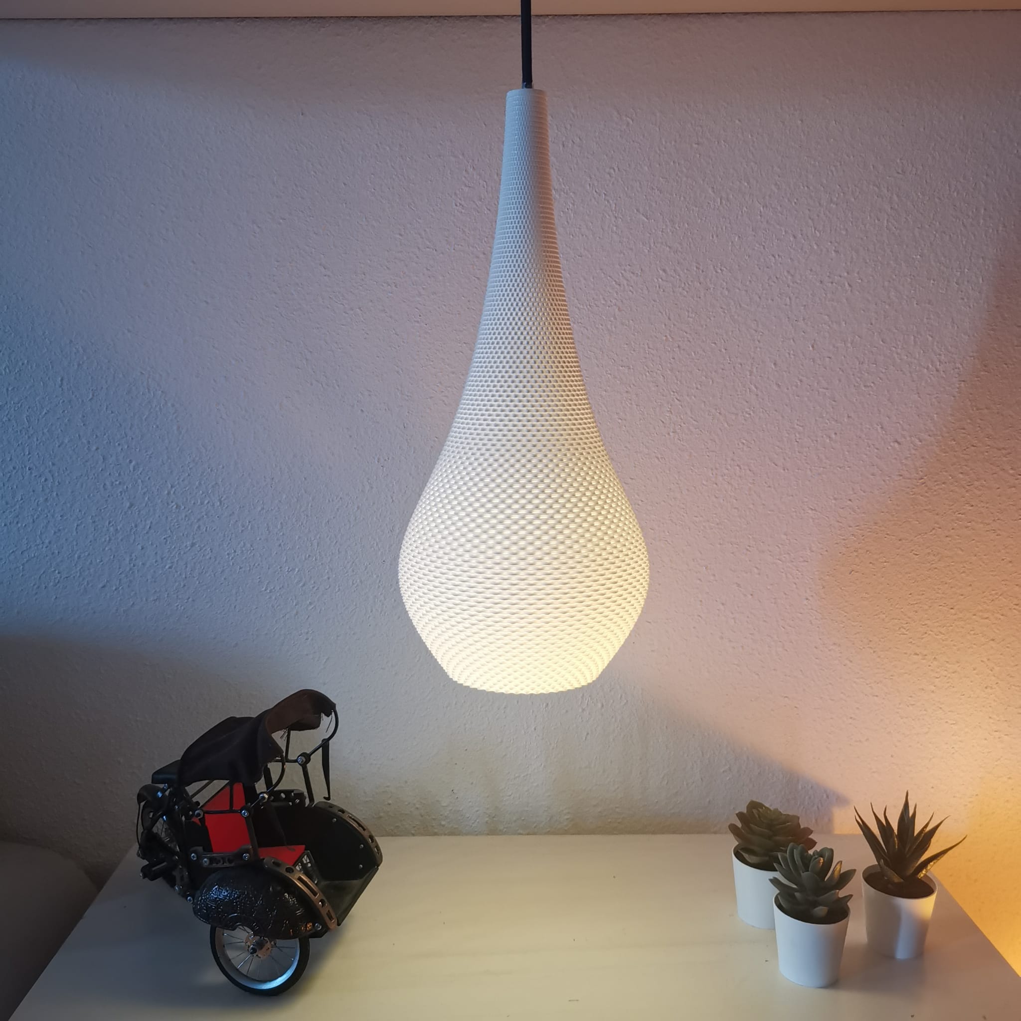 White textured pendant light hanging above a small model motorcycle and three potted plants on a white surface.