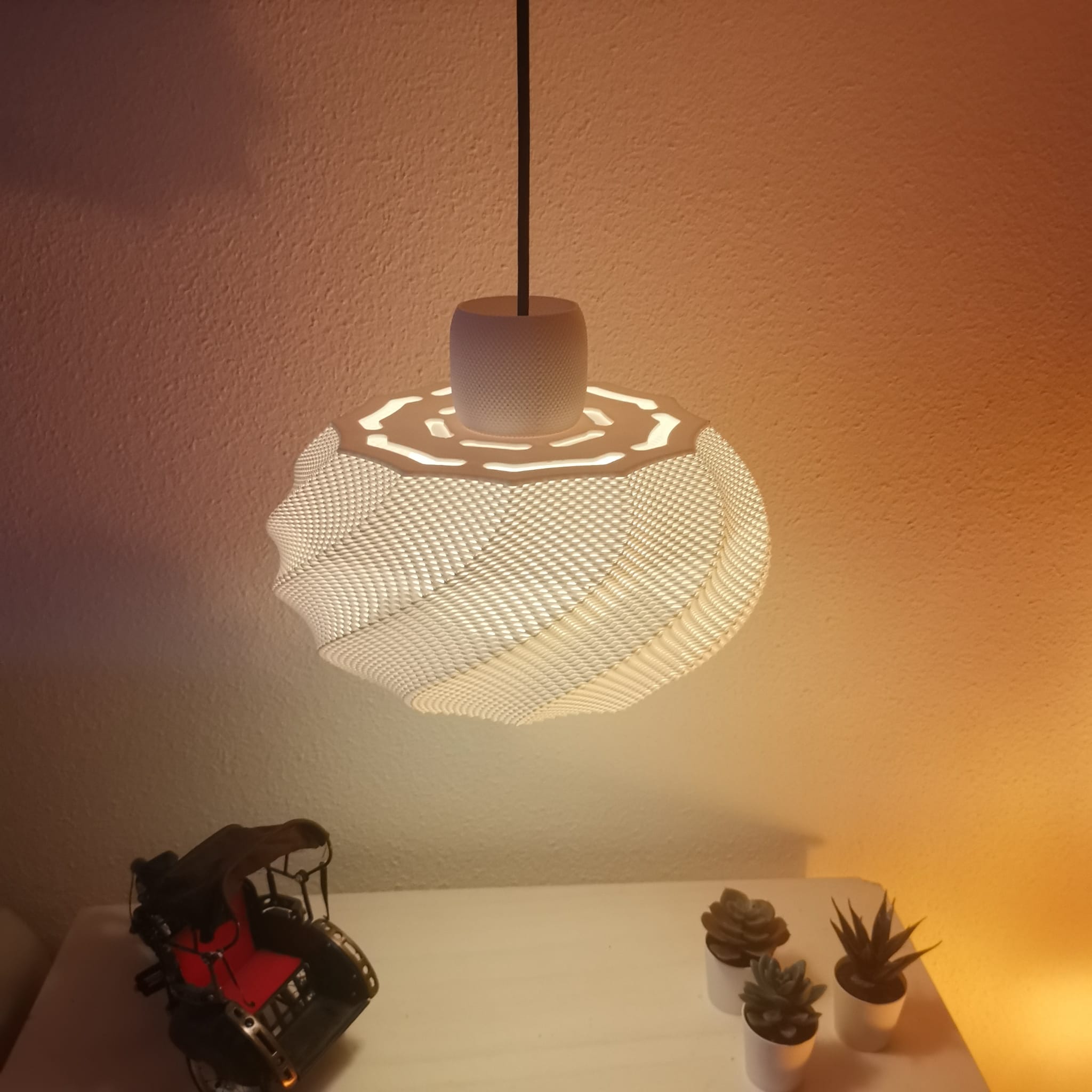 Modern pendant light fixture casting a pattern on a wall with a small model car and plants on a surface below.