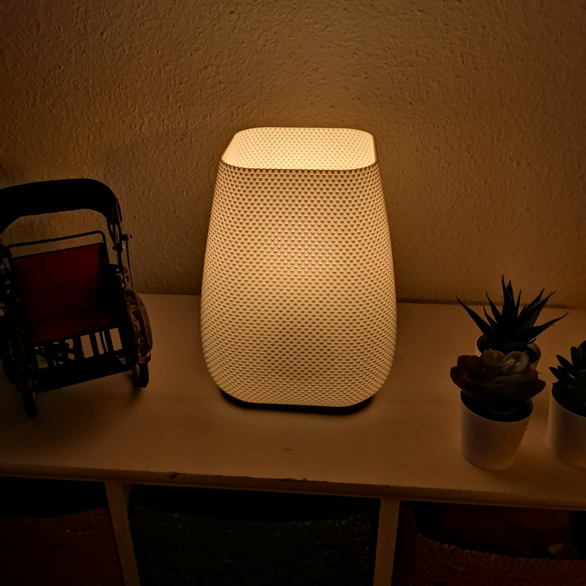 Modern textured lamp glowing softly on shelf beside decorative rickshaw and small potted succulents, warm minimalist home lighting.