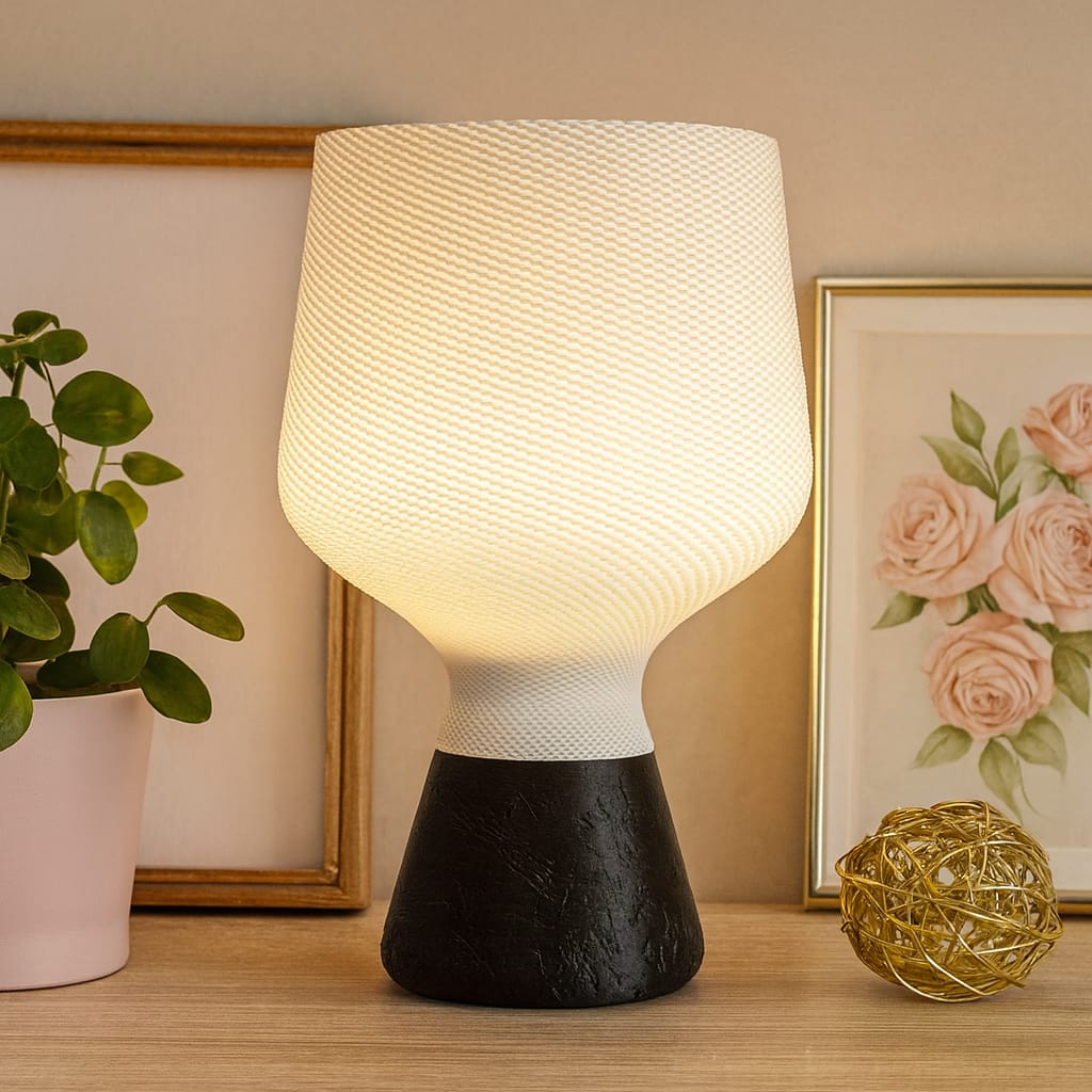 Table lamp with a textured white shade and black base on a wooden surface, with a plant and artwork in the background.