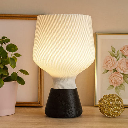 Table lamp with a textured white shade and black base on a wooden surface, with a plant and artwork in the background.